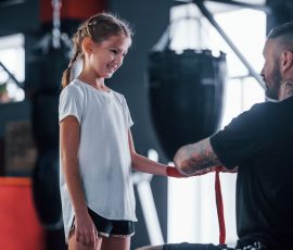 Helping to wear the bandages. Young tattooed boxing coach teaches cute little girl in the gym